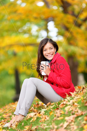Happy woman drinking coffee in fall forest / city park outdoor. Girl sitting relaxing enjoying hot drink, coffee or tea in disposable cup outside in beautiful fall foliage. Asian Caucasian female, 20s