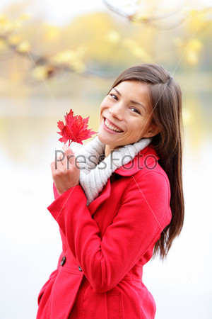 Fall girl holding red Autumn leave outside. Asian woman outdoor portrait in red seasonal autumn coat by fall forest lake. Female model smiling happy looking at camera. Mixed race Asian Caucasian girl.