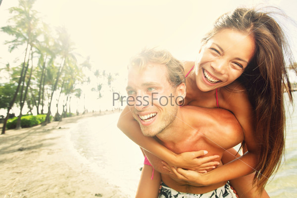 Happy Young Joyful Couple Having Beach Fun Piggybacking Laughing Together During Summer Holidays Vacation On Tropical Beach. Beautiful Energetic Fresh Interracial Multi-Ethnic Couple, Man And Woman.