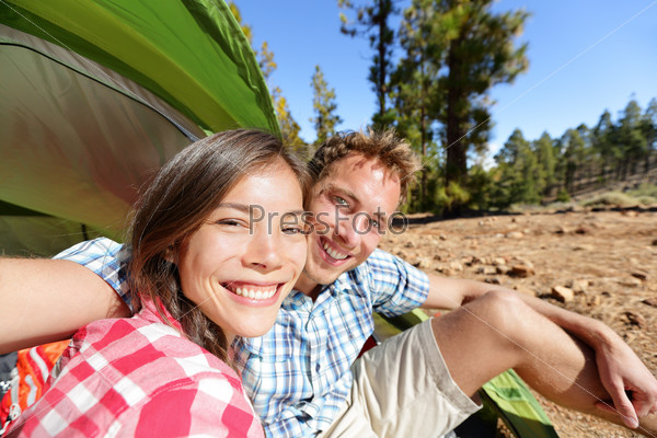 Selfie camping couple in tent taking self portrait using camera smartphone. Campers taking picture smiling happy outdoors in forest. Happy people having fun. Asian woman, Caucasian man.