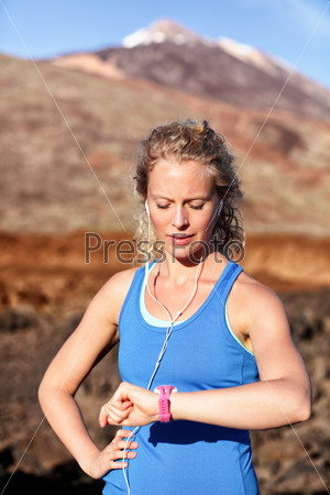 Runner looking at heart rate monitor sports smart watch after running with earphones listening to music. Female athlete checking pulse during workout run exercise outdoors in beautiful landscape.