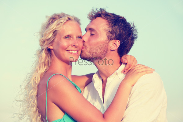 Romantic couple kissing in love enjoying sunset at beach. Young happy man kissing woman on cheek in romance on summer beach during honeymoon vacation holidays travel.