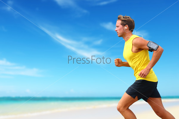Runner running listening smartphone music on beach cardio workout. Male athlete jogging on ocean beach or waterfront working out with smart phone app device and earphones in summer.