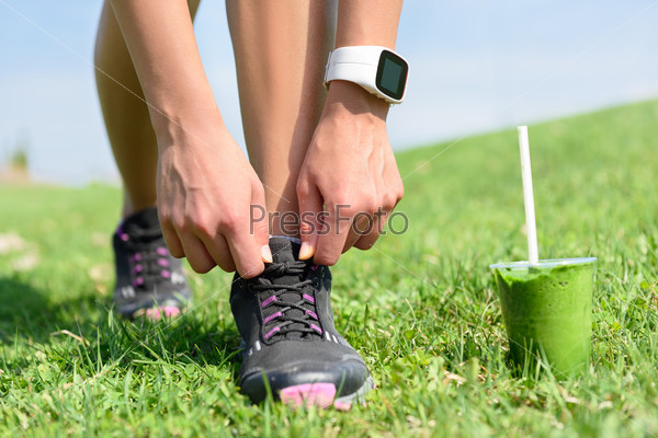 Running shoes, sports smartwatch and green smoothie. Female runner tying shoe laces in city park while drinking a healthy spinach and vegetable smoothie using smart watch heart rate monitor.