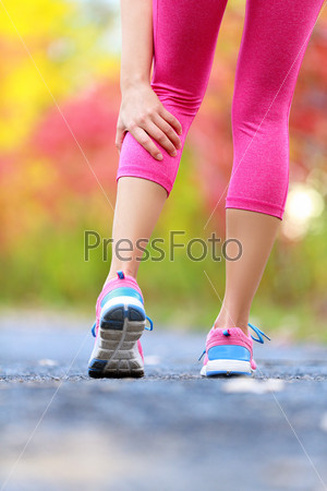 Woman running clutching calf muscle injury after spraining it while out jogging on the beach. Female athlete sport injury. Outside on forest path.