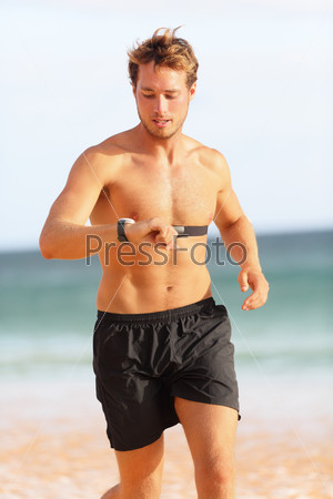 Male runner looking at heart rate monitor sports smart watch after running with earphones listening to music. Man athlete checking pulse during workout run exercise outdoors at ocean beach in summer.