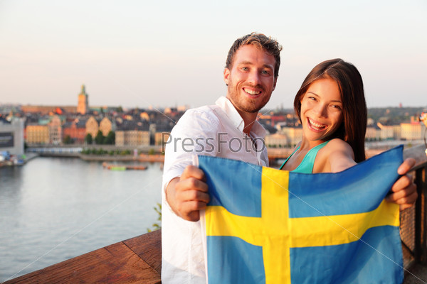 Swedish people holding Sweden flag in Stockholm. Candid fresh Scandinavian man and Asian woman looking at old town cityscape sunset view from Monteliusvagen overlooking Gamla Stan, the old city.