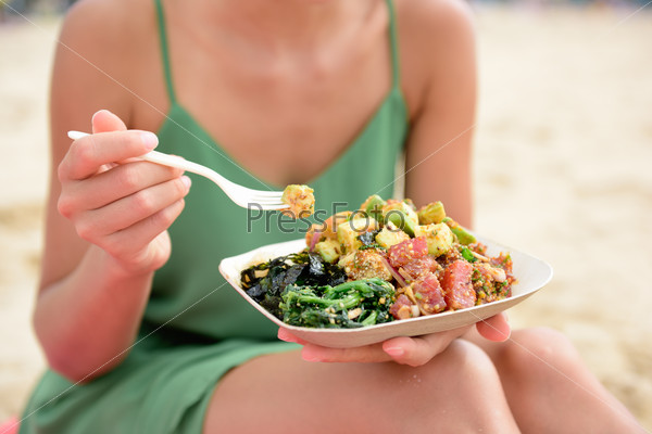 Poke bowl salad plate. A traditional local Hawaii food dish with raw marinated ahi yellowfin tuna fish. Woman sitting on beach eating lunch.