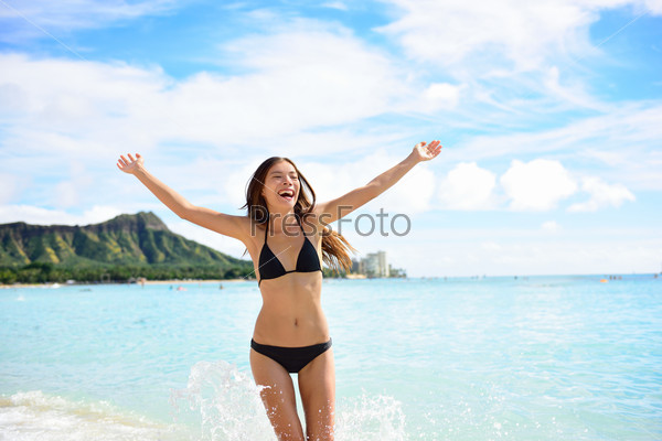 Beach fun woman happy on Hawaii vacations. Cheering Asian girl in black bikini with arms up in happiness playing and running in water on Waikiki beach, Honolulu city, Oahu island, Hawaii, USA.