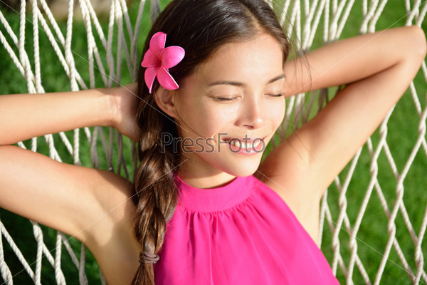 Sleeping woman relaxing on outdoor hammock in the sun tanning and having a nap in home garden or beach resort on summer holidays. Asian young adult portrait wearing pink hair flower and halter top.