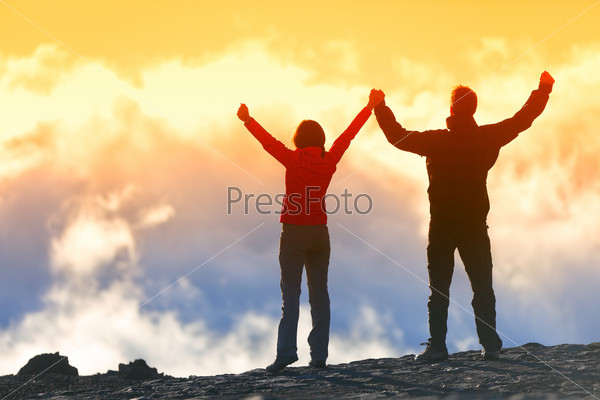 Happy winners reaching life goal - success people at summit. Business achievement concept. Two person couple together arms up in the air of happiness with accomplishment in the clouds at sunset.