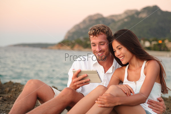 Romantic couple relaxing on beach using tablet computer at sunset. Young multiracial couple using apps on digital tablet taking pictures or reading e book looking at touch screen together.