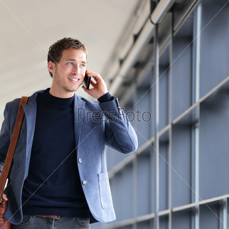 Urban business man talking on smart phone traveling walking in full body length inside in airport. Casual young businessman wearing suit jacket and shoulder bag. Handsome male model in his 20s.