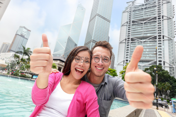 Happy multiethnic/ multicultural tourists couple showing thumbs up in satisfaction travelling in Hong Kong city, Asia. Young Asian and Caucasian adults smiling at camera - travel people