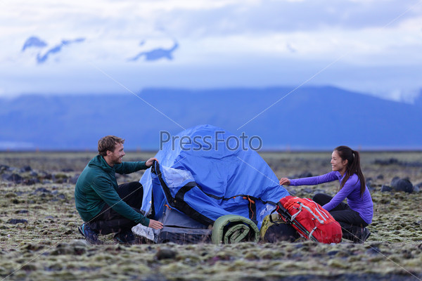Tent - people pitching tent on Iceland at dusk. Couple setting up camp for night after hiking in the wild Icelandic nature landscape. Multicultural Asian woman and Caucasian man healthy lifestyle.