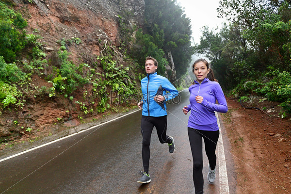 Healthy lifestyle people running on country road exercising. Runners jogging on mountain road training for marathon. Asian woman and Caucasian man wearing waterproof sports clothing for wind and rain.