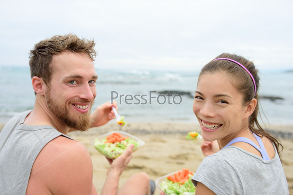 Vegan friends eating vegetarian salad meal during lunch break on beach happy looking at camera. Multiethnic group of young people, Caucasian man, Asian chinese mixed race woman in their 20s.