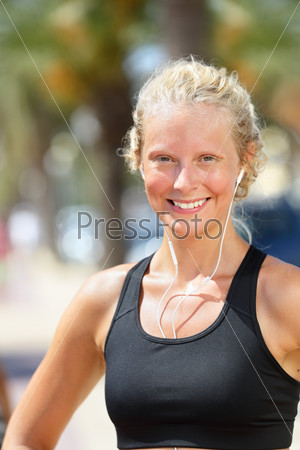 Sporty healthy woman runner listening to music. Beautiful young blond female jogger smiling at camera during run with headphones outdoors in summer. Running or fitness concept.