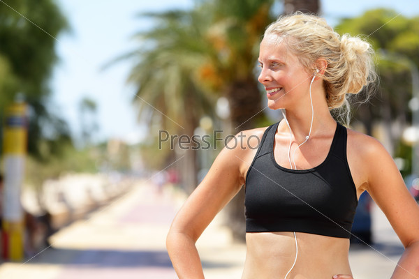 Active fitness woman in sports bra and earphones listening to music. Pretty blonde female runner looking to the side happy and motivated before going jogging and running in the city on a summer day.