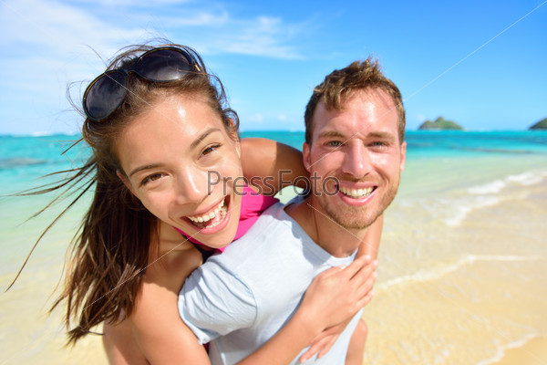 Happy beach couple in love on summer vacations on Lanikai beach, Oahu, Hawaii, USA with Mokulua Islands. Joyful Asian girl piggybacking on young Caucasian boyfriend and having fun on travel holiday.