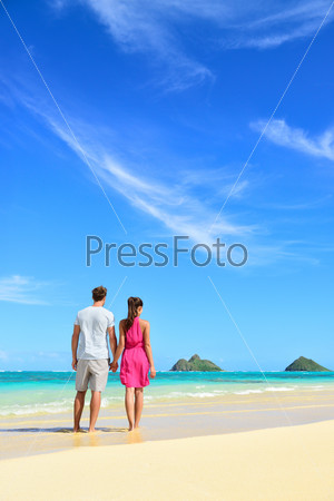 Beach vacation couple relaxing on summer holidays. Young people standing from behind holding hands looking at the ocean on Lanikai beach, Oahu, Hawaii, USA with Na Mokulua Islands.