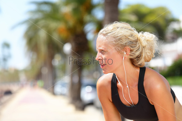 Tired running woman taking a break during run. Beautiful young blond athletic female adult resting catching her breath while jogging and listening to music with earphones and smartphone app.
