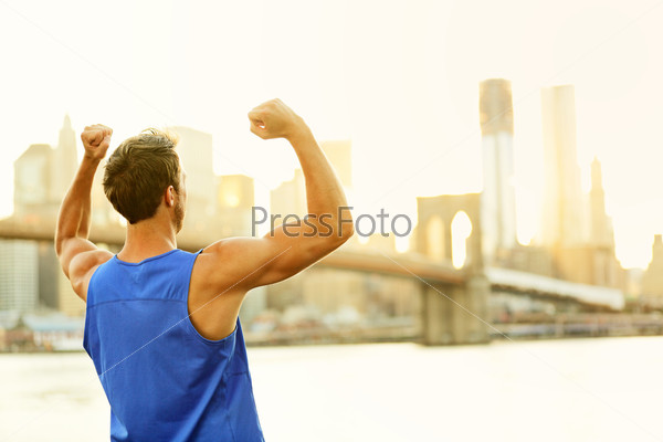 Winning cheering success fitness runner man in New York City skyline celebrating happy and flexing strong muscles after running workout training outside with Brooklyn Bridge in background.