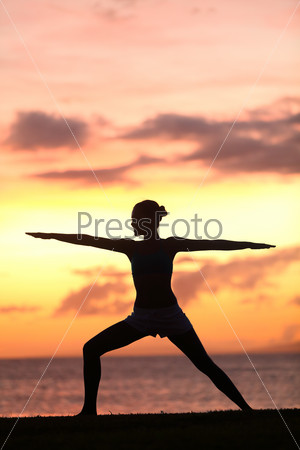 Yoga woman training and meditating in warrior pose outside by beach at sunrise or sunset. Female yoga instructor working out training in serene ocean landscape. Silhouette of woman model against sun.