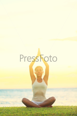 Yoga woman relaxing by sea at sunrise or sunset doing the Sukhasana, easy pose facing water. Woman meditating in beautiful ocean landscape retreat. Meditation, yoga and relaxation concept.