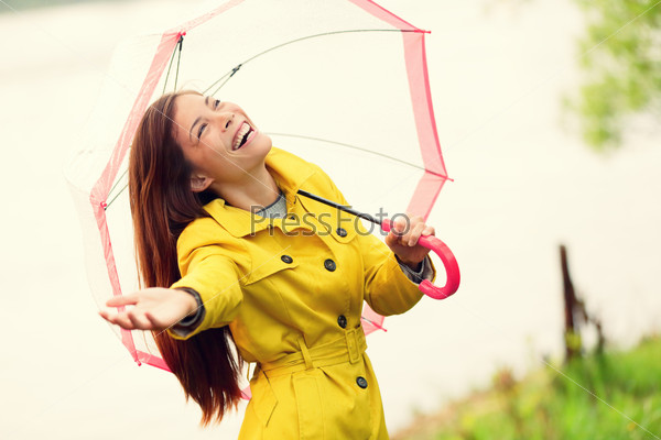 Fall woman happy after rain walking with umbrella. Female model looking up at clearing sky joyful on rainy Autumn day wearing yellow raincoat outside in nature forest by lake. Mixed race Asian girl.