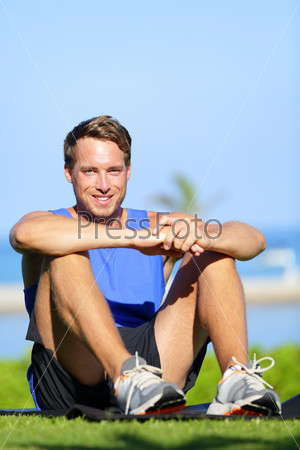 Fitness sports athlete man relaxing after training and exercising. Young male athlete resting sitting in grass after running and training exercise outside in summer. Caucasian man sports model.
