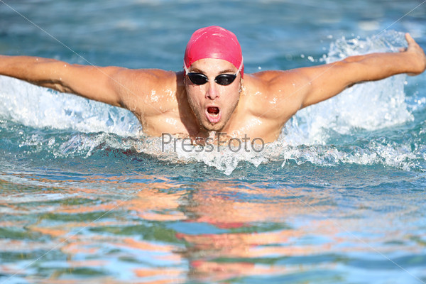 Swimmer athletic man swimming butterfly in a swimming cap approaching the camera rising up out of the water. Male athlete fitness model training outside.