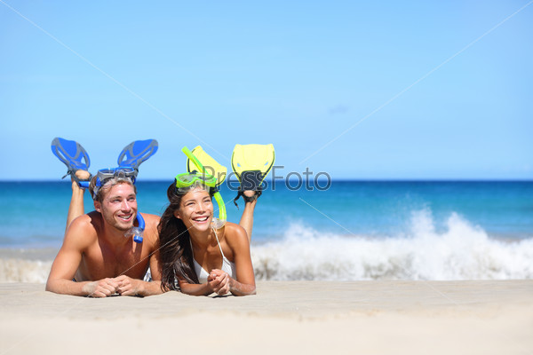 Beach travel couple having fun snorkeling. Happy young multiracial couple lying on summer beach sand with snorkel equipment looking to side at copy space after swimming with fins and mask on vacation.