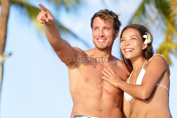 Couple on beach happy in swimwear, man pointing showing and looking at view. Beautiful young multi-ethnic couple, Asian woman and Caucasian man having fun together on summer holidays vacation travel.