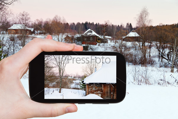 travel concept - tourist takes picture of rural landscape with snowy wooden houses in country at pink winter sunset on smartphone, Russia