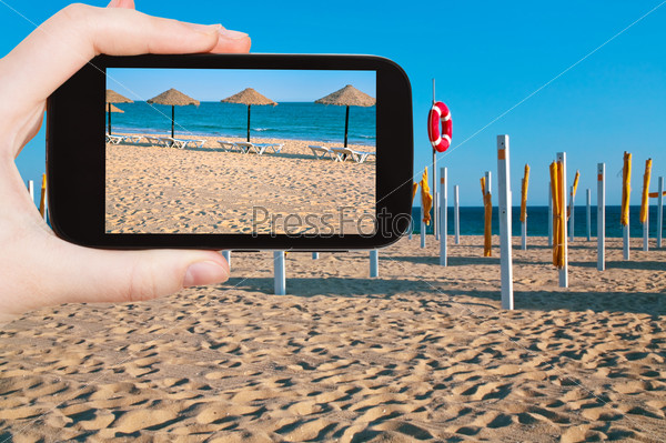travel concept - tourist taking photo of straw beach umbrellas on ocean coast, Portugal, Algarve on mobile gadget