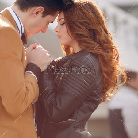 Happy couple in love,a man with dark hair in a light brown jacket and white striped shirt and a beautiful woman with long red curly hair,in a black dress,gently hugging and talking standing on the street spring city