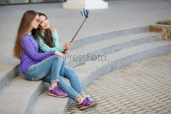 young beautiful friends students taking selfie stick picture together in town happy on sunny day. Closeup of two cheerful friends having fun and taking photos of themselves on smart phone.