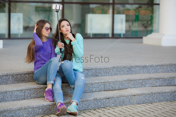 young beautiful friends students taking selfie stick picture together in town happy on sunny day. Closeup of two cheerful friends having fun and taking photos of themselves on smart phone.