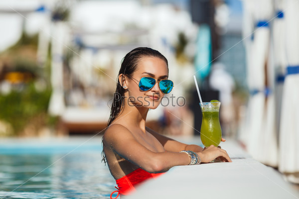 Tenned beautiful woman in orange bikini and sunlasses sitting in swimming pool with cocktail. Fashionable portrait. Elegant woman in a bikini and sunglasses