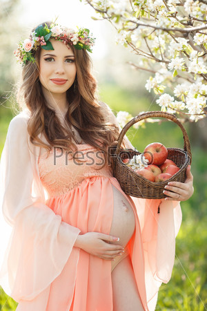 Pregnant woman posing in a green apple garden with apple in her hands. Young pregnant woman relaxing in park outdoors, healthy pregnancy. Beautiful pregnant woman holding her tummy.