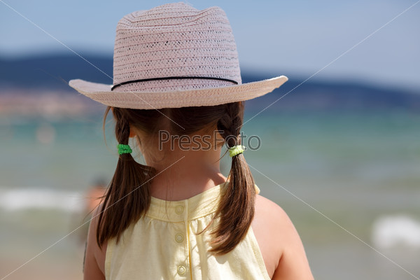 Little girl in white hat with tails looks at the sea on a bright sunny day. View from the back. Shallow depth of field. Focus on the model.