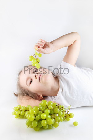 Child, a little girl lying holding a small bunch of grapes and looking into the camera. Bunch of grapes out of focus in the foreground. Vertical shot. Space for text.