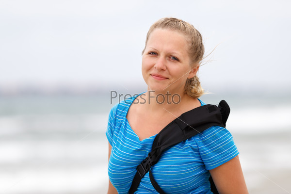 Photo of a smiling young blonde woman in a blue t-shirt with a backpack strap over his shoulder outdoors. Shallow depth of field. Focus on the model\'s face.