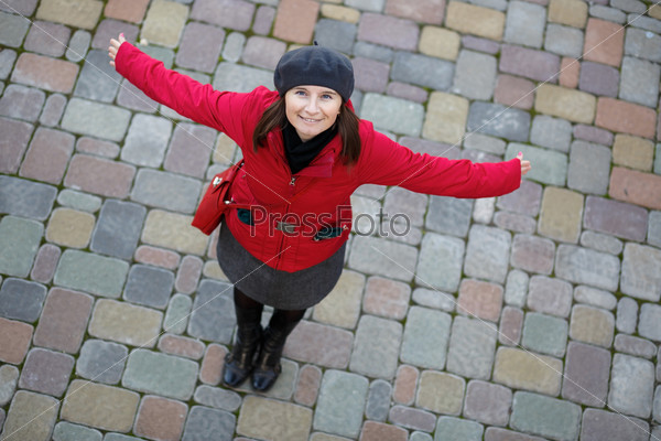Smiling woman in a red jacket and a gray beret on a background of old paving slabs. Model posing, arms to the side and looking up at the camera. Shallow depth of field. Focus on the model\'s face.