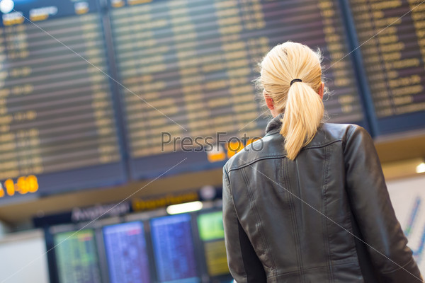 Casually dressed young stylish female traveller checking a departures board at the airport terminal hall in front of check in couters. Flight schedule display blured in the background. Focus on woman.