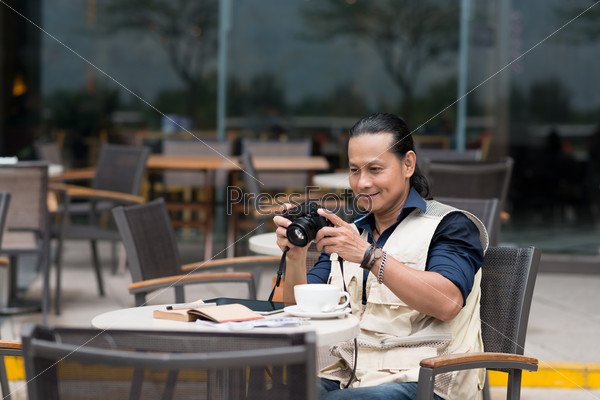 Vietnamese tourist sitting in outdoor cafe and watching pictures on his digital camera