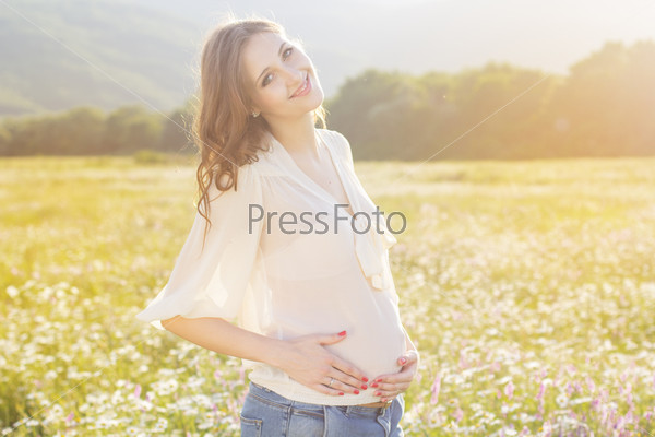 Happy pregnant woman is resting in meadow of chamomile flowers, sunset time, new life concept