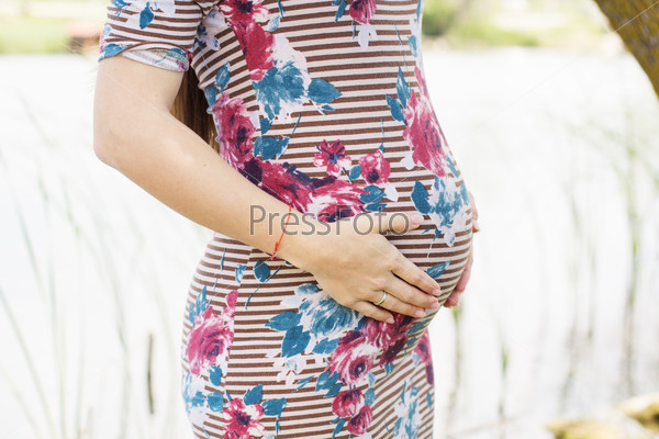 Belly of pregnant girl is wearing stripped dress is resting near blue river, summer time, pregnancy girl