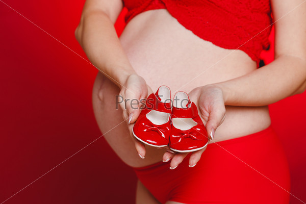 Portrait of Pregnant woman holding pair of red tiny shoes for baby on red background. Studio